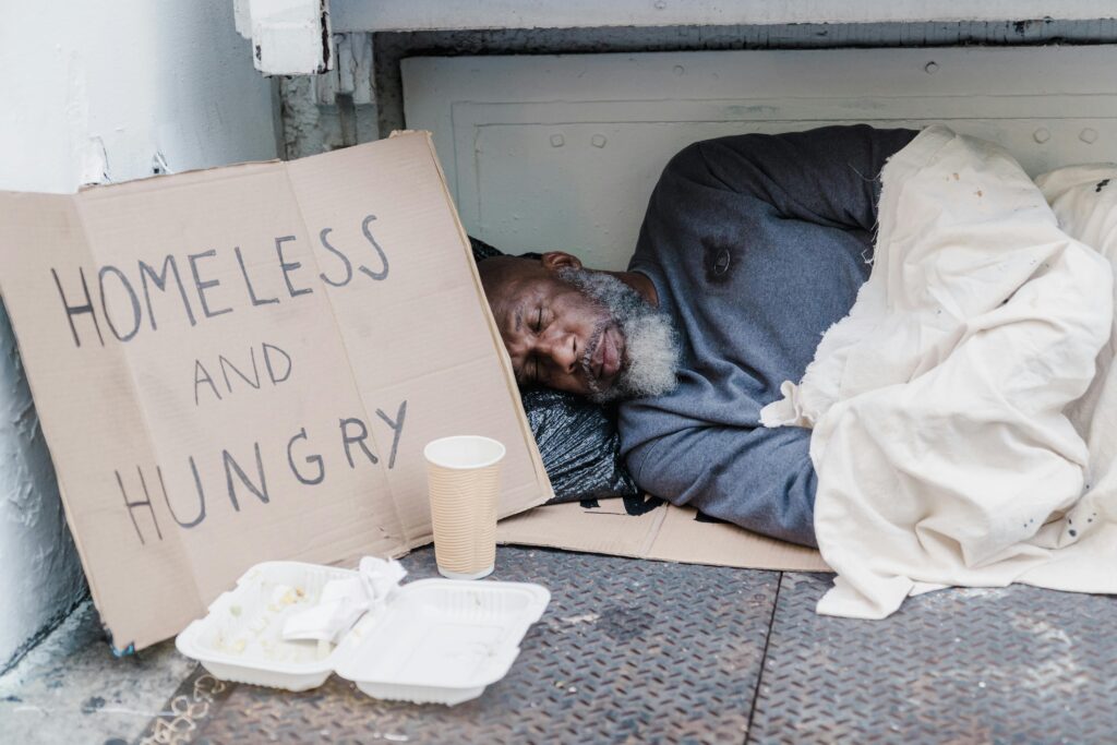 pexels-photo-9532305-9532305 Elderly homeless man sleeping on the street with a sign reading 'Homeless and Hungry'.